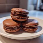Three rich, dark brown chocolate macarons stacked and arranged on a white plate.