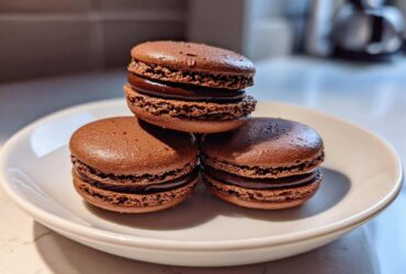 Three rich, dark brown chocolate macarons stacked and arranged on a white plate.