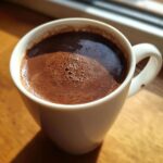Close-up of rich, dark French hot chocolate with foam in a white mug, sitting on a wooden surface.