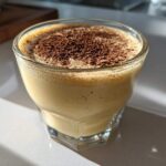 A close-up of a glass filled with creamy, frothy homemade eggnog, topped generously with ground cinnamon or nutmeg.