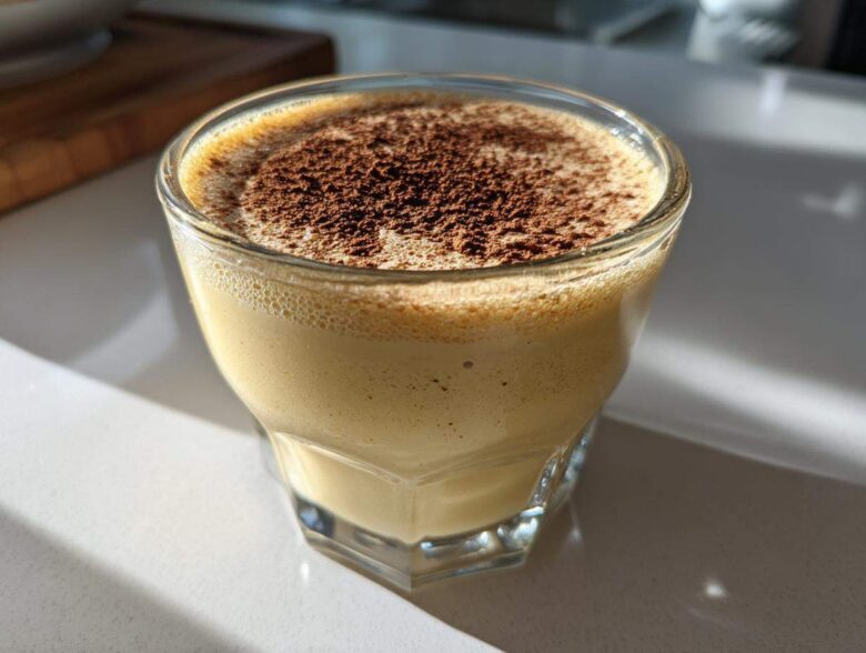 A close-up of a glass filled with creamy, frothy homemade eggnog, topped generously with ground cinnamon or nutmeg.