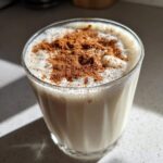 A glass of creamy, frothy coquito topped generously with ground cinnamon.