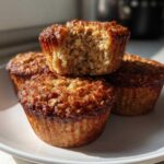 Four golden-brown banana oatmeal muffins stacked on a white plate, one with a bite taken out showing the texture.