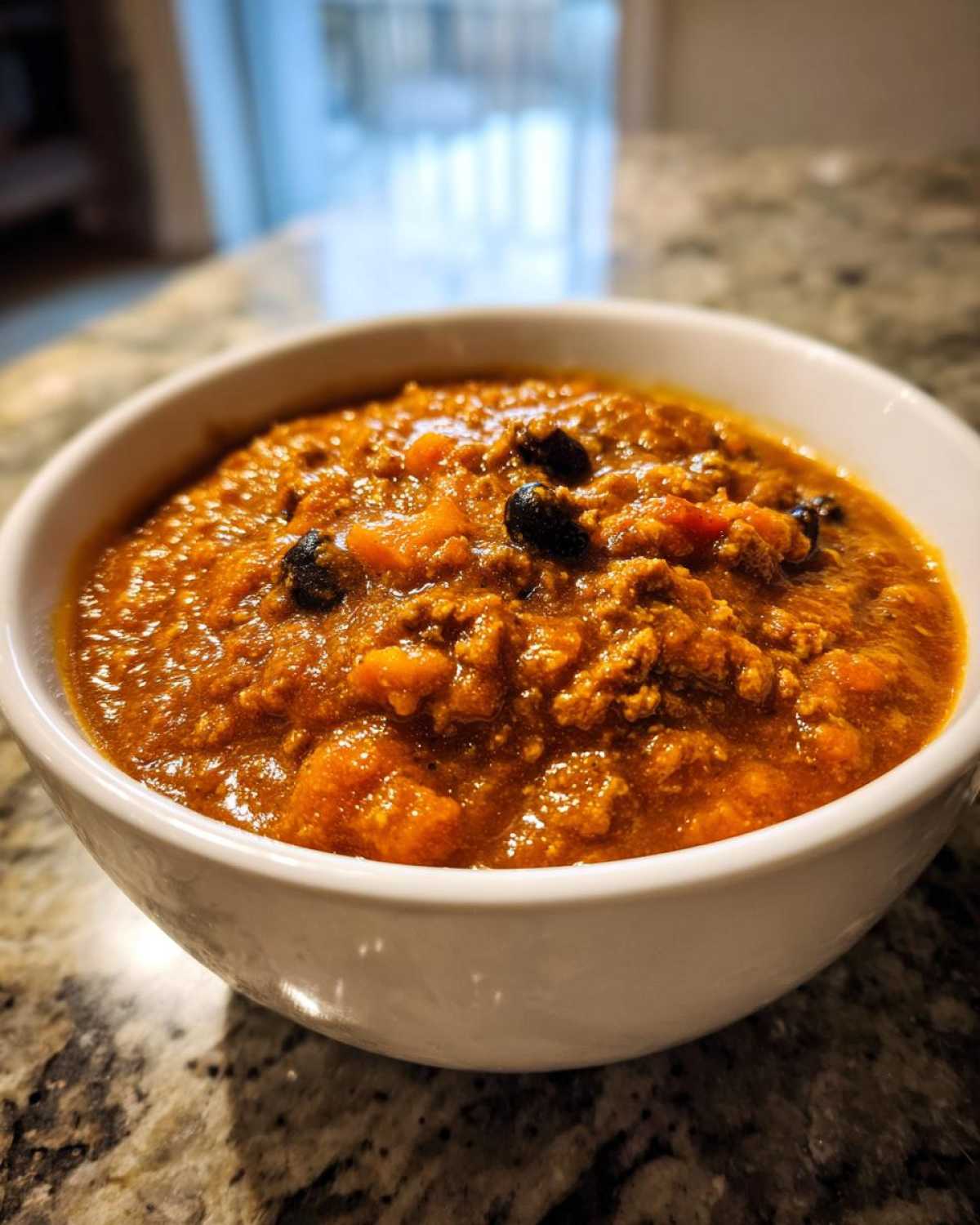 Close-up of a hearty bowl of thick, orange-red pumpkin chili with visible chunks of pumpkin and black beans.