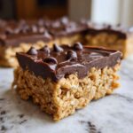 A close-up view of a homemade, chewy bar with a thick chocolate layer on top, resembling a no-bake protein bars recipe.
