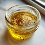 Close-up of homemade salad dressing, an emulsified mixture of golden oil, garlic, and spices, in a small glass jar.