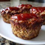 Four perfectly baked mini meatloaf portions topped with a generous dollop of bright red ketchup glaze on a white plate.