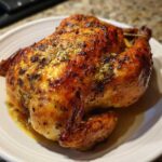 A whole, juicy roasted chicken with crispy, golden-brown skin seasoned with herbs, resting on a white plate.