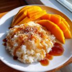 A serving of mango sticky rice topped with sweet sauce and toasted sesame seeds, garnished with fresh mango slices.