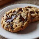 Two freshly baked vegan chocolate chip cookies with gooey, melted chocolate chunks on a white plate.