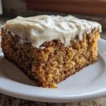 A square slice of moist applesauce cake topped with thick, creamy white frosting, served on a white plate.