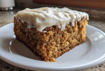 A square slice of moist applesauce cake topped with thick, creamy white frosting, served on a white plate.