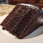 A close-up view of a moist slice of dark chocolate cake with rich, glossy frosting.