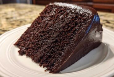 A close-up view of a moist slice of dark chocolate cake with rich, glossy frosting.
