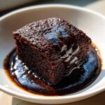 A square slice of rich, dark chocolate pudding cake swimming in a glossy chocolate sauce.