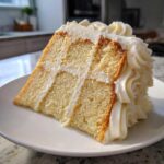 Close-up of a tall slice of moist vanilla cake recipe layered with thick white frosting.