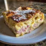 A thick, golden slice of Monte Cristo breakfast casserole filled with ham and creamy cheese, dusted with powdered sugar.