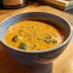Close-up of a bowl of rich, orange broccoli cheddar soup with visible pieces of broccoli florets.