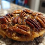 Close-up of a single, glossy pecan pie cookie topped with whole pecans and sticky caramel filling.