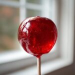 Close-up of a perfectly coated, shiny red candy apples on a wooden stick against a bright window background.