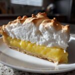 A tall slice of lemon meringue pie featuring a thick, toasted meringue topping over bright yellow lemon filling.
