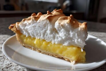 A tall slice of lemon meringue pie featuring a thick, toasted meringue topping over bright yellow lemon filling.