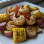 A close-up of a plated sheet pan shrimp boil featuring seasoned shrimp, corn on the cob pieces, and red potatoes.