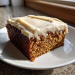 A moist slice of dark brown applesauce cake topped with thick, creamy white frosting, sitting on a white plate.