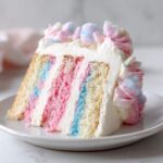 A tall slice of cotton candy cake showing colorful pink and blue layers alternating with white frosting, topped with cotton candy.