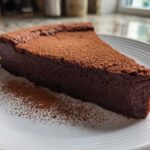 A thick slice of dark, dense flourless chocolate torte dusted heavily with cocoa powder on a white plate.