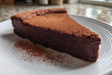A thick slice of dark, dense flourless chocolate torte dusted heavily with cocoa powder on a white plate.