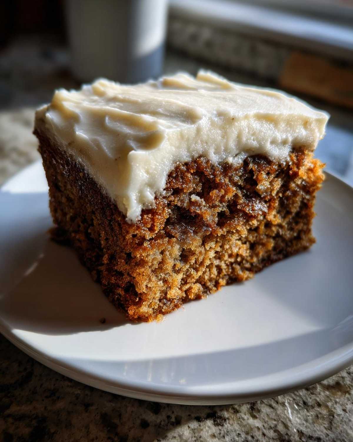 A moist, dark slice of applesauce cake topped with thick white cream cheese frosting, sitting on a white plate.