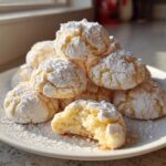 A pile of soft, crinkle-topped cool whip cookies heavily dusted with powdered sugar on a white plate.