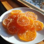 Close-up of glistening candied orange slices coated in sugar, resting on a white plate.