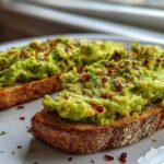 Two slices of rustic bread topped with chunky, bright green avocado toast and sprinkled with red pepper flakes.