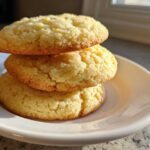 Three thick, golden brown sugar cookies stacked vertically on a small white plate, catching sunlight.