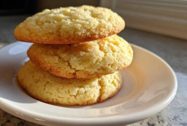 Three thick, golden brown sugar cookies stacked vertically on a small white plate, catching sunlight.