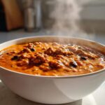 Close-up of a steaming white bowl filled with thick, rich pumpkin chili featuring ground meat and black beans.