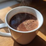 A close-up of a steaming white mug filled with rich, dark French hot chocolate, catching sunlight on a wooden surface.