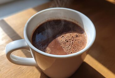 A close-up of a steaming white mug filled with rich, dark French hot chocolate, catching sunlight on a wooden surface.
