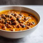 Close-up of a steaming white bowl filled with rich, orange-hued pumpkin chili containing ground meat and black beans.