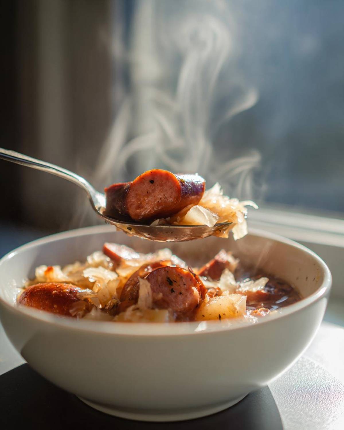 A spoonful of steaming hot sauerkraut soup, featuring sliced sausage and cabbage, being lifted from a white bowl.