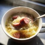 A spoonful of hot sauerkraut soup with thick slices of smoked sausage being lifted from a white bowl.