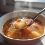 A spoonful of steaming sauerkraut soup, showing sausage, potatoes, and carrots, lifted from a white bowl.
