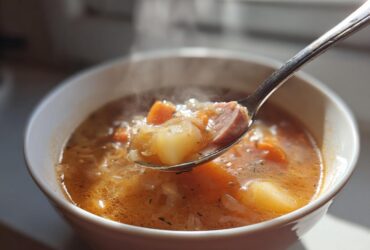 A spoonful of steaming sauerkraut soup, showing sausage, potatoes, and carrots, lifted from a white bowl.