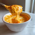 A spoonful of thick, gooey, orange beer cheese dip is lifted from a white bowl, showing long cheese pulls.