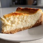 A close-up slice of creamy sugar cookie cheesecake with a golden-brown top and thick crust on a white plate.
