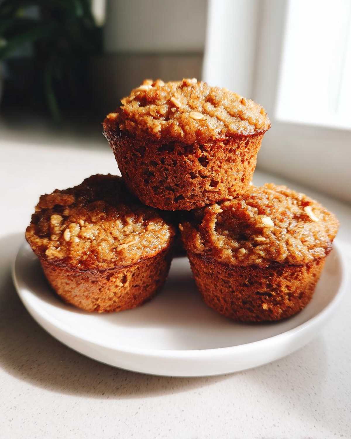 Three golden brown banana oatmeal muffins stacked on a small white plate, catching bright sunlight.