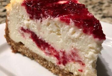 A thick slice of creamy white chocolate cheesecake featuring a bright red raspberry swirl and topping, resting on a graham cracker crust.
