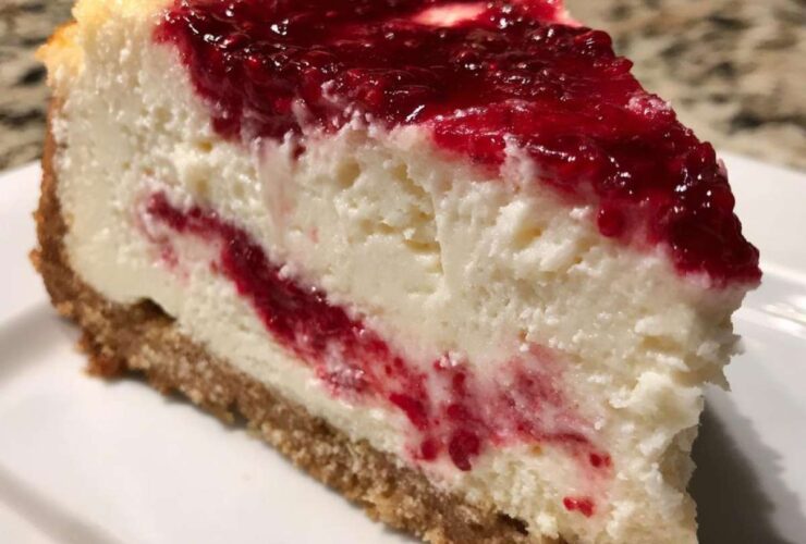 A thick slice of creamy white chocolate cheesecake featuring a bright red raspberry swirl and topping, resting on a graham cracker crust.